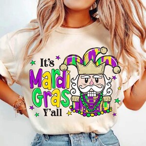 Puede incluir: Una camiseta blanca con un diseño gráfico colorido que presenta una figura de bufón con una corona y un collar de Mardi Gras morado y verde. El texto "It's Mardi Gras Y'all" está impreso en la camiseta.