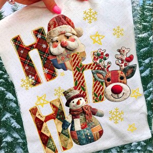 May include: White Christmas-themed decorative towel with the word "HO HO HO" in patchwork letters. Santa Claus, a reindeer, and a snowman are featured. Gold snowflakes and stars are scattered around the design.