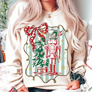 May include: Cream-colored sweatshirt featuring a festive Christmas design. The graphic includes a red and white polka-dot bow, a green Christmas tree, and a red nutcracker soldier. The design is framed with a green border and set against a striped background.