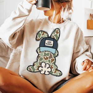 May include: A white sweatshirt featuring a camouflage bunny wearing a blue and black baseball cap with the text "egg hunter".
