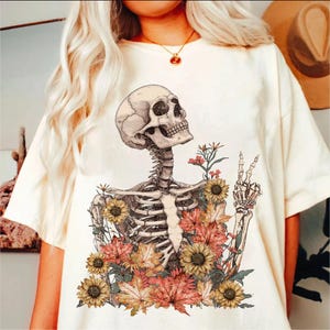 May include: A white t-shirt with a graphic of a skeleton surrounded by flowers. The skeleton is making a peace sign with its hand. The flowers are in shades of orange, yellow, and red.