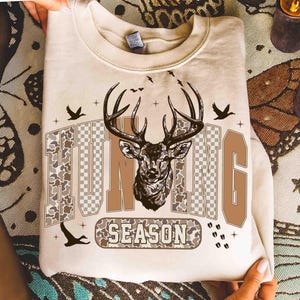 K&ouml;nnte beinhalten: Beiges Sweatshirt mit einem Hirschkopf-Motiv und den Worten "HUNTING SEASON" in Braun und Camouflage-Farben. Das Design umfasst Vogelsilhouetten und Kreuze, wodurch eine &Auml;sthetik zum Thema Jagd entsteht.