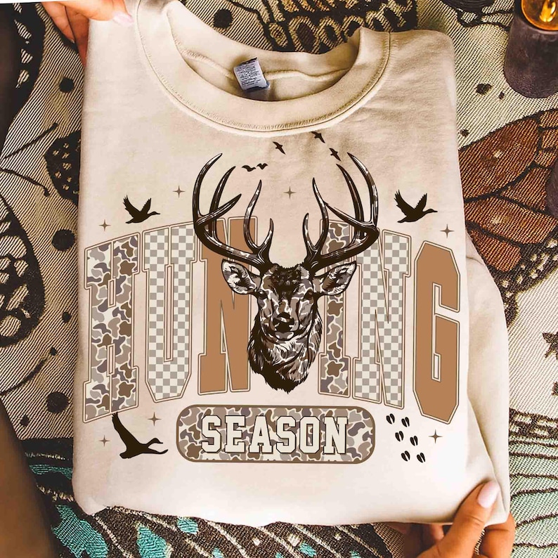 K&ouml;nnte beinhalten: Beiges Sweatshirt mit einem Hirschkopf-Motiv und den Worten "HUNTING SEASON" in Braun und Camouflage-Farben. Das Design umfasst Vogelsilhouetten und Kreuze, wodurch eine &Auml;sthetik zum Thema Jagd entsteht.