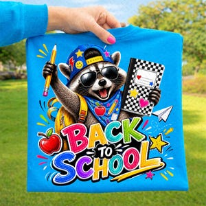 Puede incluir: Una camiseta azul brillante con un gráfico de un mapache que lleva una gorra azul y gafas de sol. El mapache sostiene un lápiz amarillo y un cuaderno a cuadros blanco y negro. El texto dice BACK TO SCHOOL con una manzana roja y estrellas.