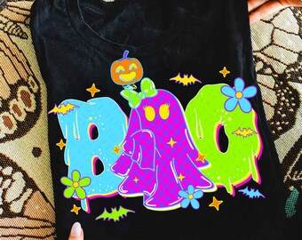 Puede incluir: Camiseta negra con la palabra "BOO" colorida en azul, rosa y verde, con un diseño de fantasma y calabaza. Incluye murciélagos, estrellas y flores, ideal para Halloween.