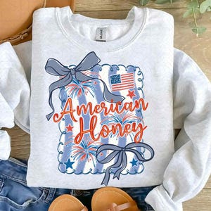 May include: A light grey sweatshirt with a patriotic design. The design features the text "American Honey" in red and orange script, with a blue and white striped background, fireworks, stars, and an American flag. Two blue bows are also included.
