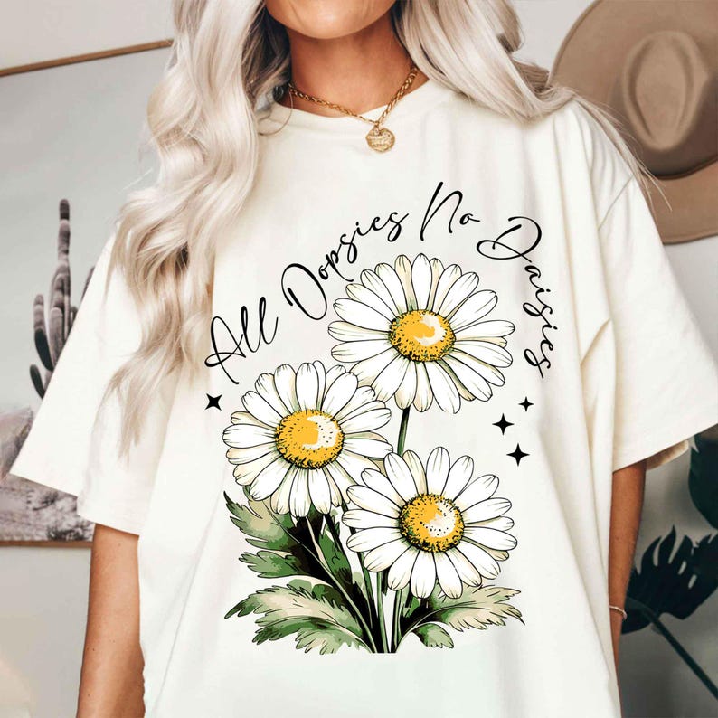 May include: White t-shirt featuring a graphic of three white daisies with yellow centres and green leaves. The text "All Daisies No Daisies" is written in black cursive.