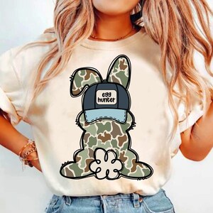 May include: White t-shirt featuring a cartoon camouflage bunny wearing a blue and black baseball cap that says "egg hunter".