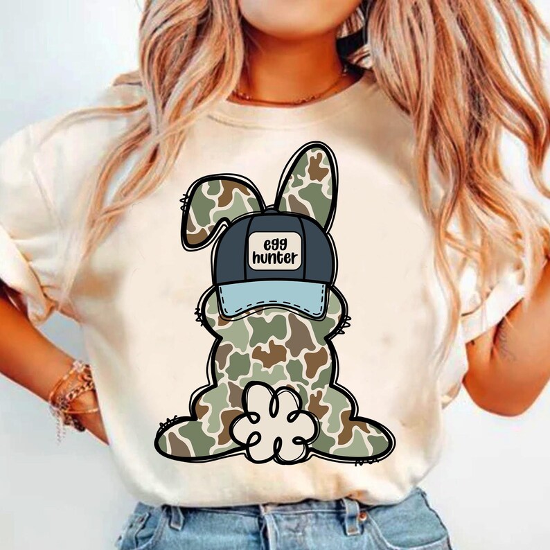 May include: White t-shirt featuring a cartoon camouflage bunny wearing a blue and black baseball cap that says "egg hunter".