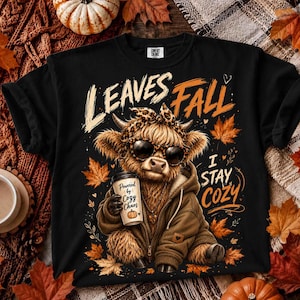 Può includere: Una t-shirt nera con la grafica di una mucca Highland con occhiali da sole, fascia leopardata e felpa marrone. La mucca tiene in mano un bicchiere di caffè con la scritta Powered by Cozy Chaos. Il testo recita Leaves Fall I Stay Cozy.