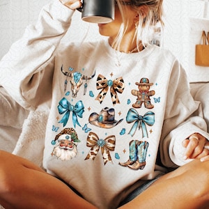 Puede incluir: Una sudadera blanca con una variedad de ilustraciones azules y marrones. Las ilustraciones incluyen una calavera con gemas turquesas, un sombrero de vaquero, una bota de vaquero, un lazo y una figura de Papá Noel.