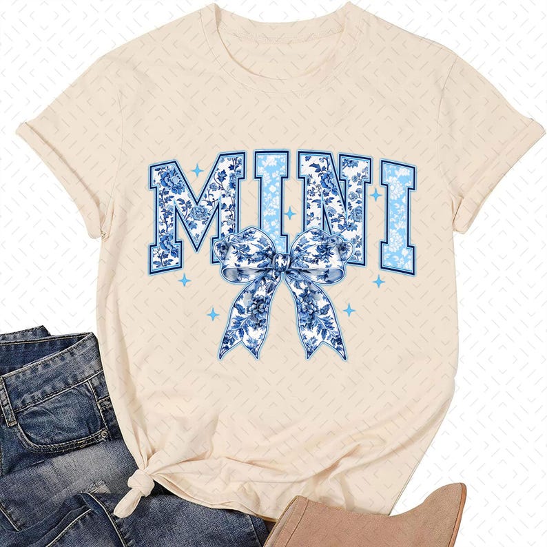 May include: A cream coloured t-shirt with the word "MINI" printed in blue and white floral patterned letters. A blue and white floral patterned bow is centred below the word.