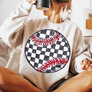 May include: Cream-colored sweatshirt featuring a baseball design. The baseball graphic has a black and white checkered pattern with red stitching. The sweatshirt is a casual, comfortable style.