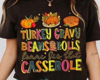 May include: A black t-shirt with a colorful Thanksgiving-themed graphic. The design includes a pumpkin, turkey, pie, and the words "TURKEY GRAVY BEANS AND ROLLS lemme see that CASSEROLE" in a vibrant, eye-catching font.