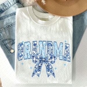 May include: White t-shirt featuring a blue and white floral design that reads "Grandma" with a blue and white floral bow.