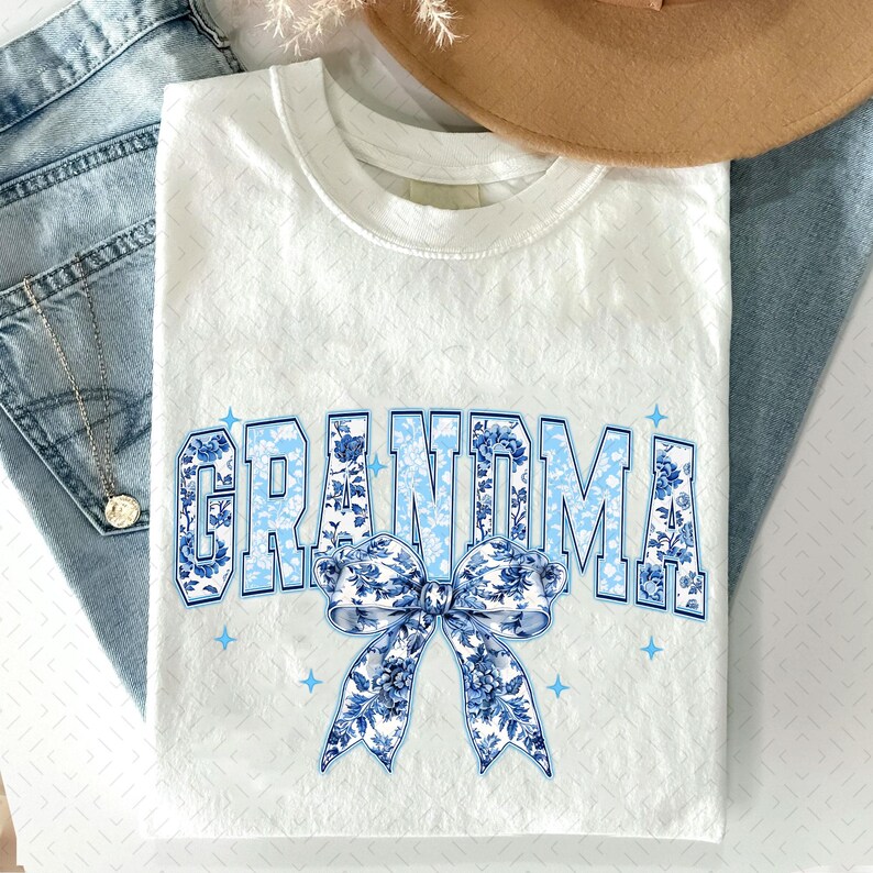 May include: White t-shirt featuring a blue and white floral design that reads "Grandma" with a blue and white floral bow.