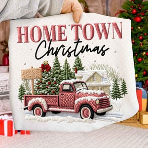 Puede incluir: Cojín bordado con las palabras "HOME TOWN Christmas" en rojo y negro. El diseño presenta una camioneta roja que transporta un árbol de Navidad, un letrero y una casa nevada, todo sobre un paisaje nevado.