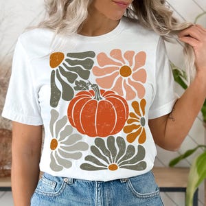 May include: White t-shirt featuring a fall-themed graphic. The design includes an orange pumpkin and stylized flowers in shades of orange, gray, and peach. The shirt is a casual, everyday wear item.