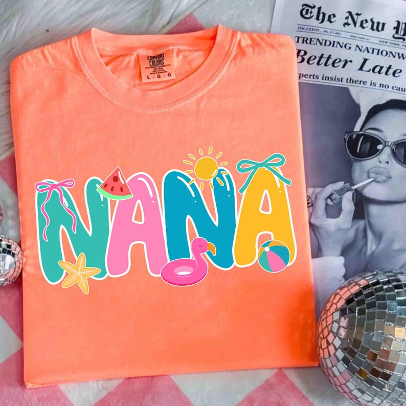 May include: Coral-colored t-shirt with the word "NANA" in colorful, playful letters. The design includes a watermelon slice, sun, flamingo float, starfish, and beach ball. The shirt has a Comfort Colors tag.