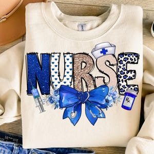 May include: A cream-colored sweatshirt with the word "NURSE" in blue, gold, and white glitter letters. The letters are decorated with blue hearts, leopard print, and a blue bow with a stethoscope. A syringe, a blue flower, and a bottle with a prescription label are also included in the design.