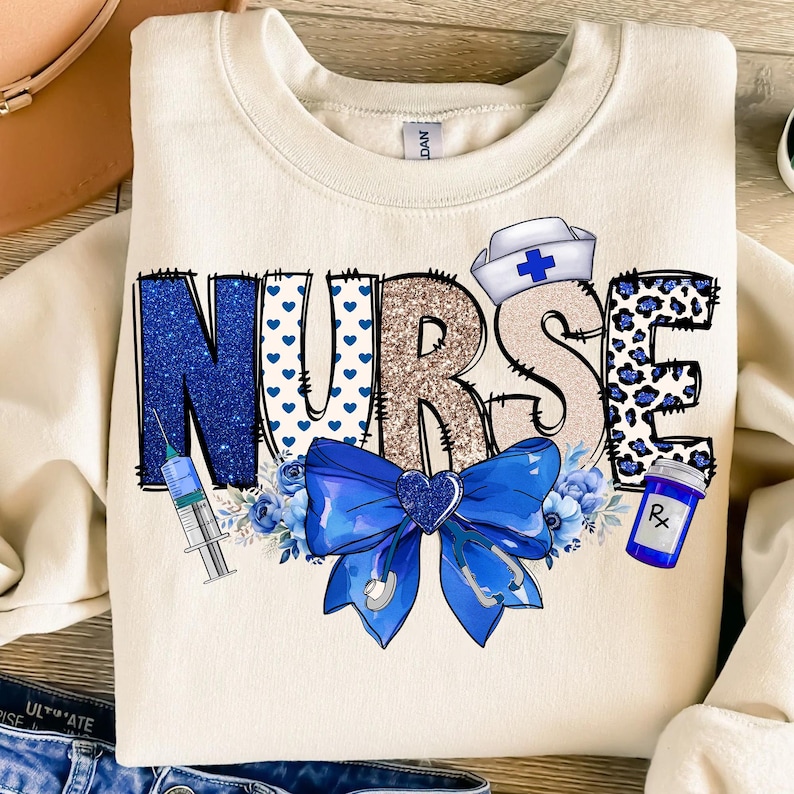 May include: A cream-colored sweatshirt with the word "NURSE" in blue, gold, and white glitter letters. The letters are decorated with blue hearts, leopard print, and a blue bow with a stethoscope. A syringe, a blue flower, and a bottle with a prescription label are also included in the design.