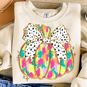 May include: A beige sweatshirt with a colorful pumpkin design. The pumpkin has a black and white polka dot bow and is outlined in gold glitter. The pumpkin is filled with a colorful abstract pattern.