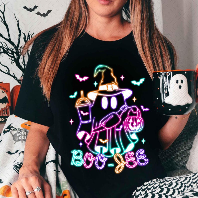May include: A black t-shirt featuring a neon pink, blue, and green graphic of a ghost wearing a witch hat and holding a pumpkin and a drink. The text "Boo-Jee" is written below the ghost in neon colours.