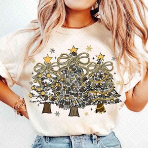 Può includere: T-shirt color crema con grafica di tre alberi di Natale con motivi mimetici. Ogni albero è adornato con un fiocco dorato, una stella e ornamenti. Il design include fiocchi di neve e la scritta "cozy".