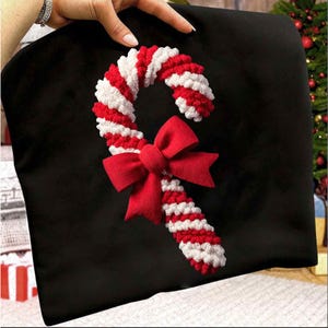 May include: Black pillow cover featuring a large, red and white candy cane design with a red bow. The candy cane is made of a textured, knitted material. A festive holiday decoration.