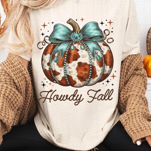 Puede incluir: Una camiseta blanca con una calabaza de estampado de vaca marr&oacute;n y blanca con un lazo turquesa y pedrer&iacute;a. El texto "Howdy Fall" est&aacute; escrito en letras marrones con forma de cuerda debajo de la calabaza.