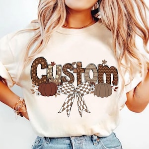 Könnte beinhalten: Ein weißes T-Shirt mit einem braunen und weißen Design, das das Wort "Custom" mit Kürbissen und einer Schleife zeigt.