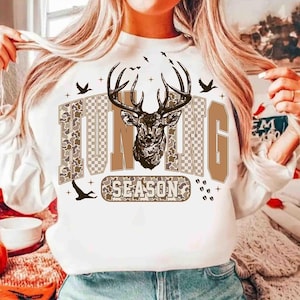 K&ouml;nnte beinhalten: Wei&szlig;es Sweatshirt mit dem Schriftzug "HUNTING SEASON" in Braun- und Camouflage-Design. Eine detaillierte Illustration eines Hirschkopfes mit Geweih befindet sich &uuml;ber dem Wort "SEASON". Mehrere V&ouml;gel fliegen dar&uuml;ber.