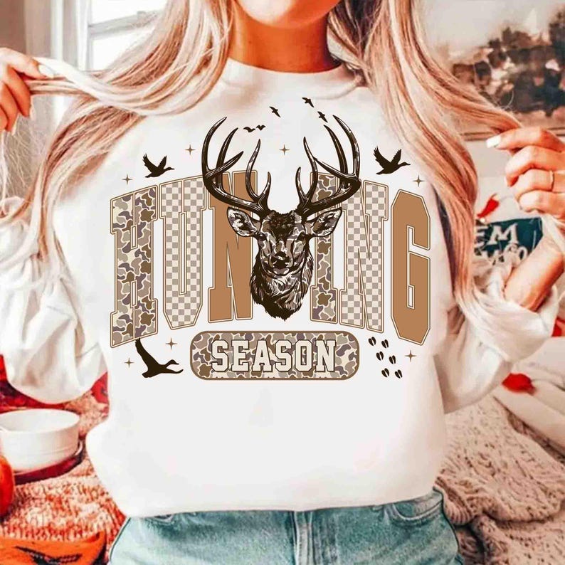 K&ouml;nnte beinhalten: Wei&szlig;es Sweatshirt mit dem Schriftzug "HUNTING SEASON" in Braun- und Camouflage-Design. Eine detaillierte Illustration eines Hirschkopfes mit Geweih befindet sich &uuml;ber dem Wort "SEASON". Mehrere V&ouml;gel fliegen dar&uuml;ber.