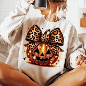 Può includere: Una felpa color crema con un design dettagliato di zucca. La zucca è arancione con un volto intagliato e un fiocco leopardato. Il design include accenti scintillanti e un piccolo pipistrello nero, perfetto per Halloween.