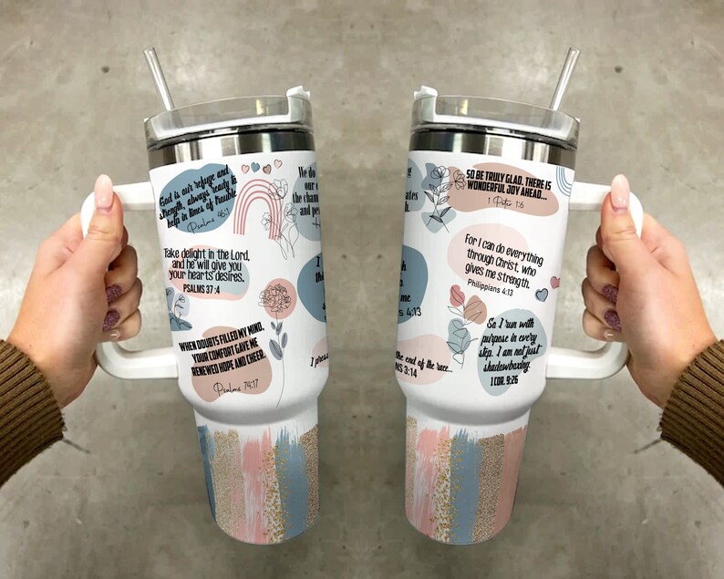 K&ouml;nnte beinhalten: Ein wei&szlig;er Becher mit Henkel und Strohhalm. Der Becher ist mit verschiedenen inspirierenden Zitaten und Designs verziert, darunter Blumen, Herzen und Pinselstriche in Rosa, Blau und Gold. Die Zitate beinhalten "Gott ist unsere Zuflucht und St&auml;rke, immer bereit, in Zeiten der Not zu helfen." und "Freut euch also wahrhaft. Es gibt eine wunderbare Freude vor euch."
