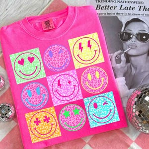 Könnte beinhalten: Leuchtend pinkes T-Shirt mit einem Raster von Smiley-Gesichtern. Jedes Smiley-Gesicht hat ein einzigartiges Muster und Farbe, einschließlich Herzen, Sterne und Blitze. Das Shirt besteht aus einem weichen Material und hat einen klassischen Rundhalsausschnitt.