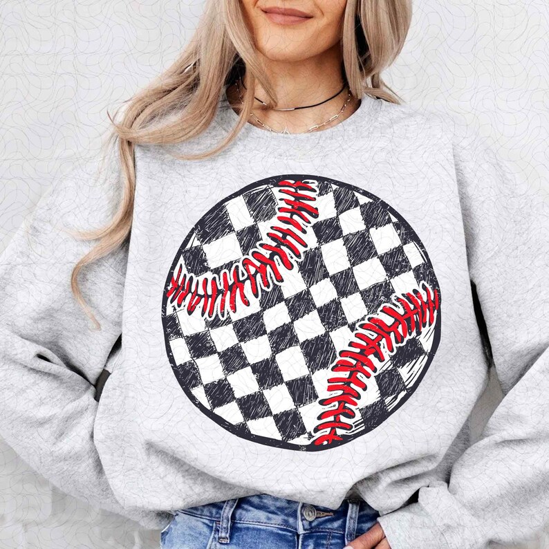 May include: A light gray sweatshirt featuring a baseball design. The baseball graphic has a black and white checkered pattern with red stitching. The design is circular and centered on the sweatshirt.
