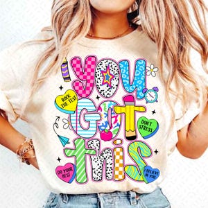 以下が含まれることがあります： 「YOU GOT THIS」と書かれたカラフルなグラフィックが特徴のクリーム色のTシャツ。デザインには、ハート、星、鉛筆、「ROCK THE TEST」、「DON'T STRESS」、「DO YOUR BEST」、「I BELIEVE IN YOU」というフレーズが含まれています。