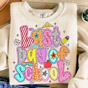 Pode incluir: Uma camisa de moletom bege com um gr&aacute;fico colorido que diz "&Uacute;ltimo dia de aula". O gr&aacute;fico &eacute; feito de diferentes formas e padr&otilde;es, incluindo estrelas, listras, bolinhas e padr&otilde;es xadrez.