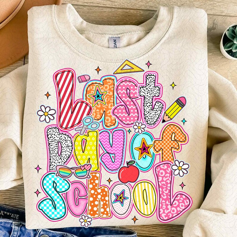 Pode incluir: Uma camisa de moletom bege com um gr&aacute;fico colorido que diz "&Uacute;ltimo dia de aula". O gr&aacute;fico &eacute; feito de diferentes formas e padr&otilde;es, incluindo estrelas, listras, bolinhas e padr&otilde;es xadrez.