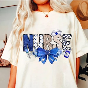 May include: White t-shirt with the word "NURSE" in blue, gold, and white letters. The letters are decorated with hearts, leopard print, and a blue bow. A syringe, a bottle of pills, and a nurse's hat are also included in the design.