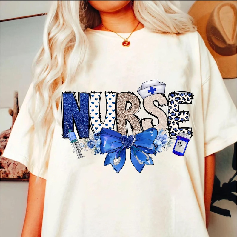 May include: White t-shirt with the word "NURSE" in blue, gold, and white letters. The letters are decorated with hearts, leopard print, and a blue bow. A syringe, a bottle of pills, and a nurse's hat are also included in the design.