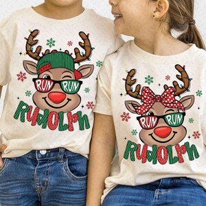 May include: Two cream-colored t-shirts with a reindeer graphic. The reindeer has sunglasses that say "RUN RUN" and a red nose. The word "Rudolph" is written below. One reindeer wears a green hat, the other a red bow.