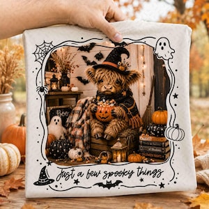 May include: A white fabric item featuring a black line-art border with spiderwebs, ghosts, and bats. The centre displays a fluffy brown highland cow wearing a witch hat and scarf, holding a jack-o'-lantern. Text reads: Just a few spooky things.