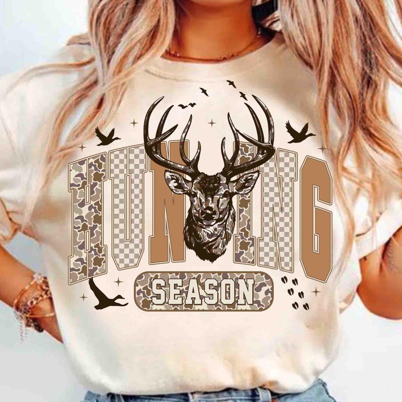 K&ouml;nnte beinhalten: Cremefarbenes T-Shirt mit einem Hirsch-Motiv und den Worten "HUNTING SEASON" in braunen und Camouflage-Buchstaben. Das Design beinhaltet Vogelsilhouetten und dekorative Elemente. Ein l&auml;ssiges T-Shirt zum Thema Outdoor.