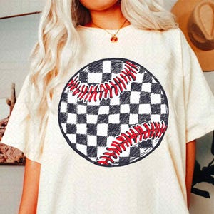 May include: Cream-colored t-shirt featuring a baseball graphic. The baseball design is a black and white checkered pattern with red stitching. The shirt has short sleeves and a crew neck.