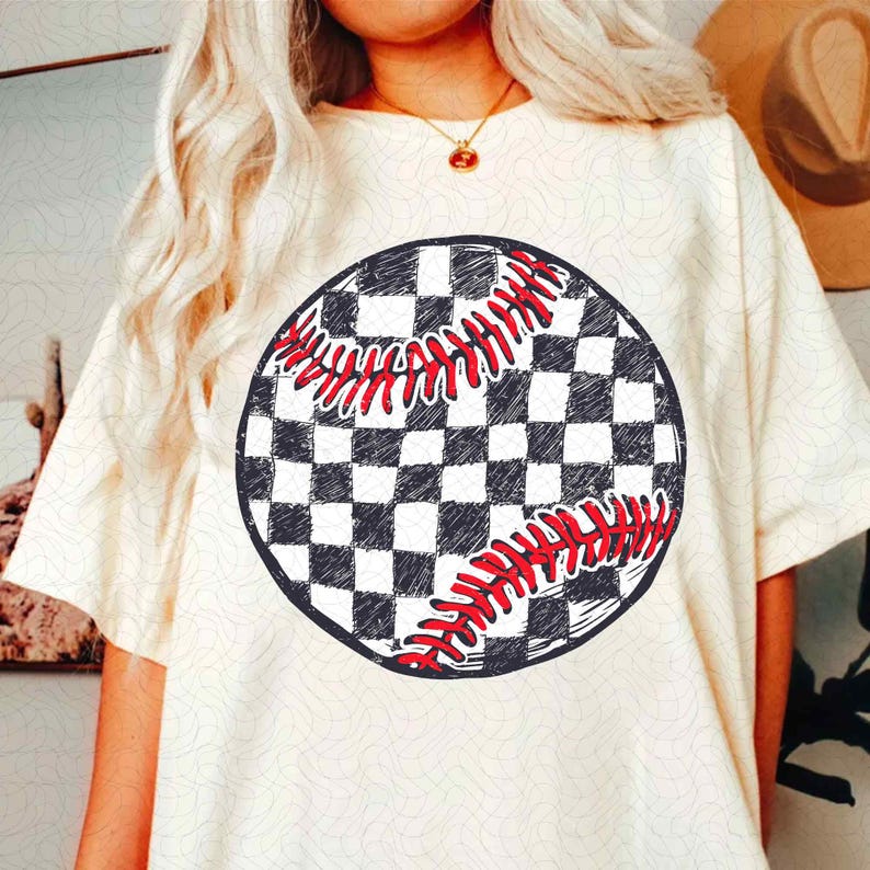May include: Cream-colored t-shirt featuring a baseball graphic. The baseball design is a black and white checkered pattern with red stitching. The shirt has short sleeves and a crew neck.