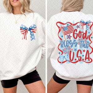 May include: A white long-sleeve sweatshirt featuring a small graphic of a red, white, and blue patriotic bow on the front left chest. The back displays a large design with the text God Bless The U.S.A, a waving American flag, and fireworks.