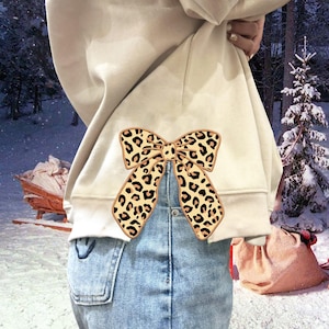 Może przedstawiać: Biała bluza z dużą kokardą w panterkę z tyłu. Kokarda jest umieszczona centralnie na plecach bluzy i zawiązana w talii. Bluza jest noszona na niebieskich dżinsach.