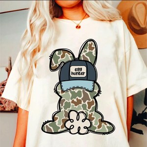 May include: White t-shirt featuring a cartoon camouflage bunny wearing a blue and white baseball cap that says "egg hunter".